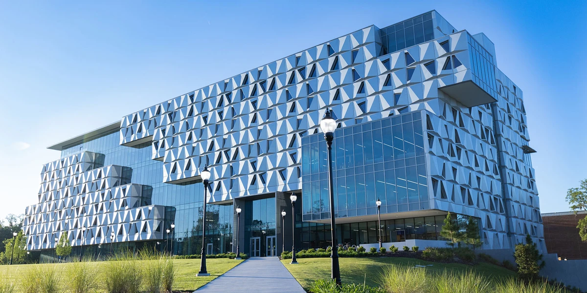 The image shows a modern, multi-story building with a unique geometric facade, set against a clear blue sky.